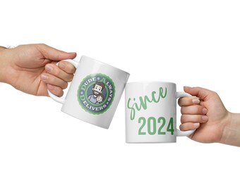 Op de afbeelding: Twee witte mokken met groene tekst en ontwerpen. Een mok zegt "Dude Always Delivers" met een cartoonafbeelding van een vader en zoon. De andere mok zegt "sinds 2024".