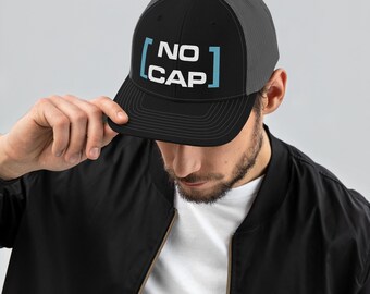 Op de afbeelding: Zwarte en grijze truckerpet met de tekst "NO CAP" in wit en lichtblauw. De pet heeft een mesh achterkant en een gebogen klep. De persoon op de afbeelding draagt een zwarte jas.
