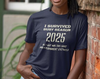 Op de afbeelding: Marineblauw T-shirt met de witte tekst "I SURVIVED BUSY SEASON 2025 ALL I GOT WAS THIS SHIRT AND A PERMANENT EYETWITCH". De persoon draagt een lichtblauwe jeans.