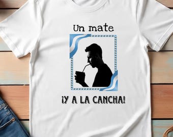 Puede incluir: Camiseta blanca con un gráfico de una persona bebiendo de una taza con pajita, enmarcado por detalles azules y blancos. Los textos "Un mate" y "¡Y A LA CANCHA!" están impresos encima y debajo del gráfico.