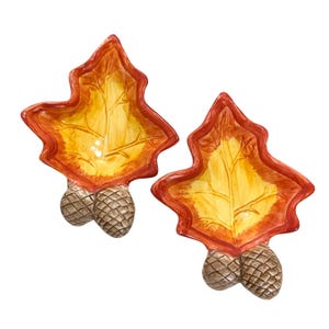 May include: Two ceramic leaf-shaped serving dishes with acorn-shaped bases. The dishes are decorated with a yellow and orange leaf design.