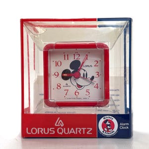 May include: A red Lorus Quartz alarm clock featuring Mickey Mouse in a clear plastic box. The clock has a white face with black numerals and hands. The box has red and blue accents and the words "Mickey & Friends" and "Alarm Clock".