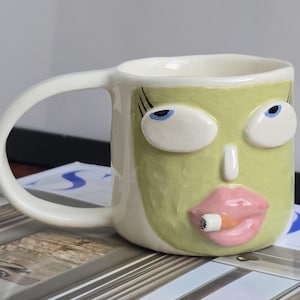 Puede incluir: Una taza de cerámica con exterior color crema y un rostro verde claro esculpido en el frente. El rostro presenta grandes ojos blancos con pupilas azules, largas pestañas negras, una nariz pequeña y labios rosados gruesos sosteniendo un cigarrillo.