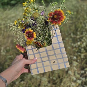 May include: A flat, rectangular ceramic vase with a beige base and a blue grid pattern, containing a bouquet of wildflowers including red and yellow blanket flowers, small white yarrow, and purple blossoms, with a small dark heart detail.