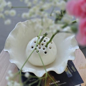 Puede incluir: Un florero de cerámica blanca con múltiples aberturas circulares se encuentra dentro de un cuenco blanco a juego con bordes ondulados. El cuenco está colocado sobre un libro negro con el texto TASCHEN y Bibliotheca Universalis, conteniendo tallos.