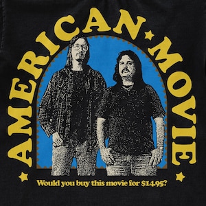 May include: A black t-shirt with a yellow and brown graphic of two men in front of a blue and white arch. The text "AMERICAN MOVIE" is written in a yellow circle around the men. The text "Would you buy this movie for $14.95?" is written below the graphic.
