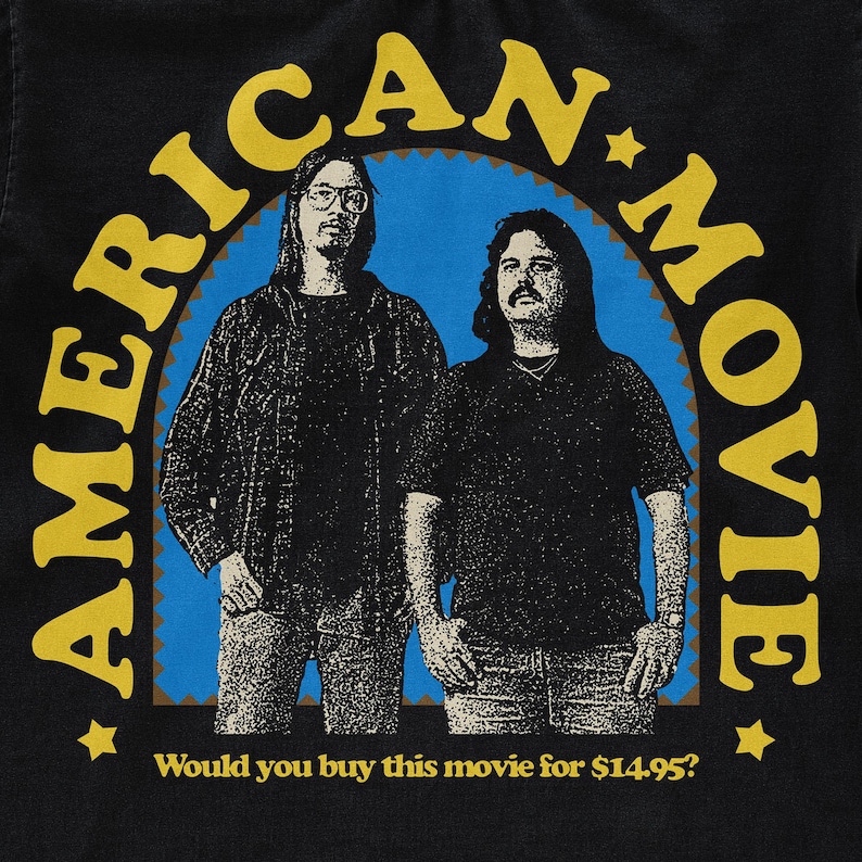 May include: A black t-shirt with a yellow and brown graphic of two men in front of a blue and white arch. The text "AMERICAN MOVIE" is written in a yellow circle around the men. The text "Would you buy this movie for $14.95?" is written below the graphic.
