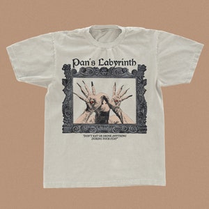Puede incluir: Una camiseta gris claro con un gráfico de estilo vintage de un hombre pálido con ojos rojos y las manos extendidas en un marco negro adornado. El texto "Pan's Labyrinth" está por encima de la imagen y "Don't eat or drink anything during your stay" está debajo de la imagen.