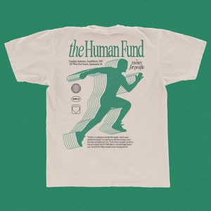 Puede incluir: Una camiseta beige con un estampado gráfico verde de una persona corriendo. El texto "the Human Fund" está impreso en verde en la parte superior de la camiseta. El texto "Vandelay Industries, Established, 1997 129 West 81st Street, Apartment 5A" está impreso en verde debajo del gráfico. El texto "money forpeople" está impreso en verde a la derecha del gráfico. El texto "I think I could be a philanthropist a kickass philanthropist! I would have all this money and people would love me. Then they would come to meand beg! And I felt like it, I would help them out. And then they would owe me big time" está impreso en verde en la parte inferior de la camiseta.