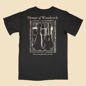 Op de afbeelding: Zwart T-shirt met een witte grafische print van een vrouw in een jurk die in een vensterframe staat. De tekst "House of Woodcock" en "Kiss me, my girl, before I'm sick." is op het shirt gedrukt.