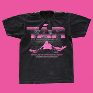 Può includere: Maglietta nera con una grafica rosa della parola "TÁR" e una persona con le braccia distese. Il testo "YOU WANT TO DANCE THE MASQUE?" è sotto la grafica.