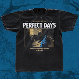 May include: A black short-sleeve t-shirt featuring the white text PERFECT DAYS, the phrase A WIM WENDERS FILM, and the years 20 and 23. Below is a rectangular graphic of a person lying down, with the text SUNLIGHT FILTERING THROUGH THE FOLIAGE and 木漏れ日.