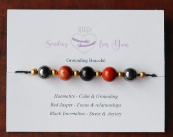 Puede incluir: Un brazalete de conexión a tierra con cuentas de turmalina negra, jaspe rojo y hematites. El brazalete está sobre una tarjeta blanca con el texto "HUGS Sending for You Grounding Bracelet Haematite - Calm & Grounding Red Jasper - Focus & relationships Black Tourmaline - Stress & Anxiety".