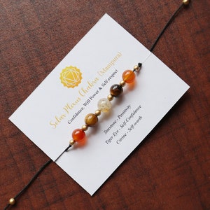 Puede incluir: Un brazalete de cuerda negra con siete cuentas. Las cuentas son de color naranja, dorado, transparente, marrón y dorado. El brazalete está sobre una tarjeta blanca con el texto "Solar Plexus Chakra (Manipura)" y descripciones de las piedras.