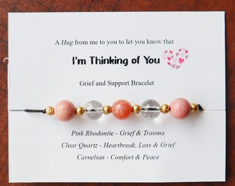 Puede incluir: Una tarjeta blanca con el texto "A Hug from me to you to let you know that I'm Thinking of You" y "Grief and Support Bracelet". Un brazalete con cuentas rosadas, transparentes y doradas se muestra en la tarjeta.