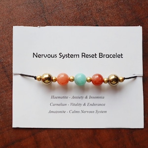 Può includere: Un braccialetto di perline con il testo "Nervous System Reset Bracelet" su una carta bianca. Il braccialetto presenta perline color oro, corallo e turchese. La carta elenca anche le proprietà delle pietre.