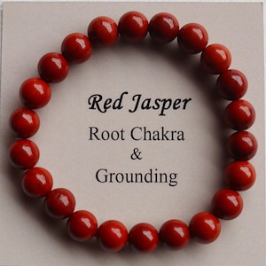 May include: A red jasper bracelet composed of round beads. The text on the card reads "Red Jasper, Root Chakra & Grounding". The bracelet is displayed on a light gray surface.