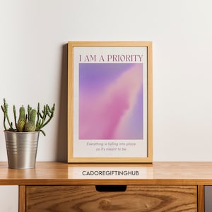 May include: A framed print with a pink and purple gradient background. The text "I AM A PRIORITY" is at the top of the print. Below the gradient is the text "Everything is falling into place as it's meant to be". The bottom of the print has the text "CADOREGIFTINGHUB".
