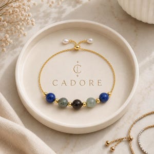 May include: A gold chain bracelet featuring five round beads in blue, green, black, and grey, accented by small gold spheres. The bracelet has an adjustable sliding closure with two small white beads at the ends, centered over the text CADORE.