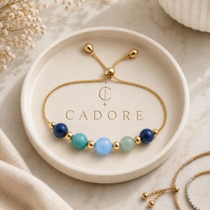 May include: A gold chain bracelet featuring five smooth, round beads in shades of dark blue, turquoise, light blue, and pale green, separated by small gold spheres. The bracelet has an adjustable sliding clasp and is displayed on a round tray.
