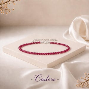 Peut inclure: Un bracelet de cheville délicat composé de petites perles facettées rose foncé disposées en cercle. Le bijou comprend un segment de chaîne en métal argenté avec un fermoir mousqueton. Le mot Cadore est écrit en lettres cursives violet foncé.
