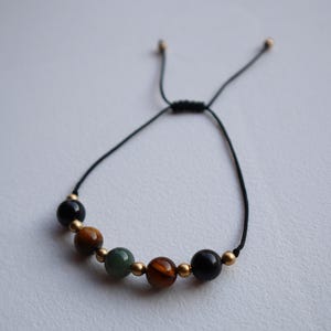 May include: A black string bracelet with gold beads and five gemstone beads. The beads are green, brown, and black.