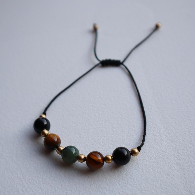 May include: A black string bracelet with gold beads and five gemstone beads. The beads are green, brown, and black.