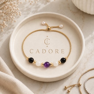 May include: A gold chain bracelet featuring a central purple amethyst bead, flanked by two white moonstone beads and two black onyx beads, accented with small gold spheres. The bracelet has an adjustable sliding closure with two small white pearls.