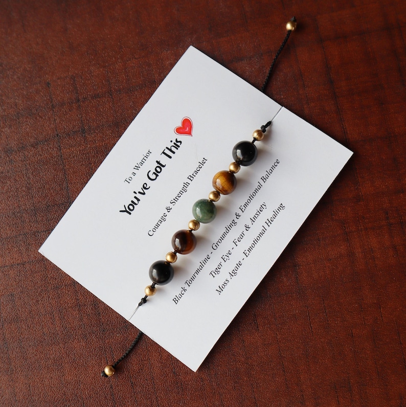 May include: A bracelet with black, green, and brown beads on a white card with the text "To a Warrior You've Got This Courage & Strength Bracelet Black Tourmaline - Grounding & Emotional Balance Tiger Eye - Fear & Anxiety Moss Agate - Emotional Healing"