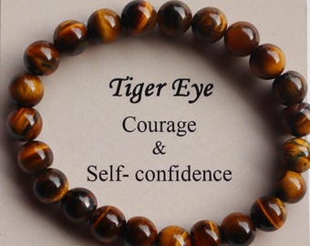 Puede incluir: Un brazalete de cuentas de ojo de tigre marrón con el texto "Tiger Eye Courage & Self-confidence" sobre un fondo beige.