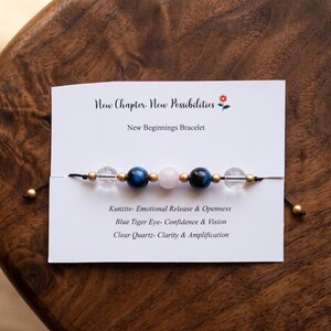 以下が含まれることがあります： New Chapter. New Possibilities、New Beginnings Bracelet、Kunzite、Blue Tiger Eye、Clear Quartzのテキストが記載された白い台紙付きのビーズブレスレット。ブレスレットには透明、青、ピンクのビーズがあしらわれています。