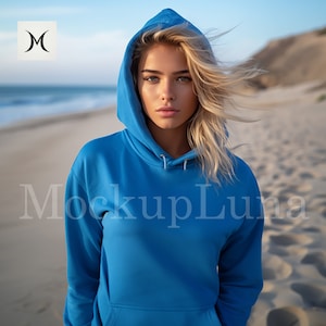 May include: A woman wearing a bright blue hooded sweatshirt with the hood up. The sweatshirt is plain and has no graphics or text. She is standing on a sandy beach with the ocean in the background.