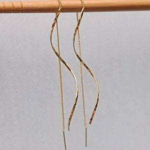 May include: A pair of gold threader earrings. Each earring has a delicate, wavy bar suspended from a fine chain. The earrings are displayed against a light background.
