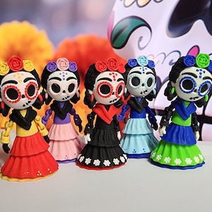 May include: Five colorful Day of the Dead figurines. Each figure has a painted skull face, flower crown, and tiered dress in red, pink, black, blue, and green. The background includes orange flowers and a decorative skull.