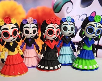 May include: Five colorful Day of the Dead figurines. Each figure has a painted skull face, flower crown, and tiered dress in red, pink, black, blue, and green. The background includes orange flowers and a decorative skull.