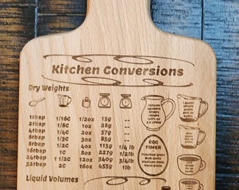 May include: A wooden chopping board with a brown stain and engraved text that reads "Kitchen Conversions". The board includes a chart of dry weights and liquid volumes with conversions for tablespoons, cups, ounces, grams, millilitres, pints, and quarts. The board also includes a chart for egg cooking times.
