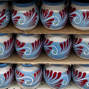 May include: A collection of small, round ceramic pots with a light gray base, blue band, and red and white floral design. The pots are arranged on cardboard shelves, showcasing a handcrafted aesthetic. The rims are painted blue, and small yellow dots accent the design.