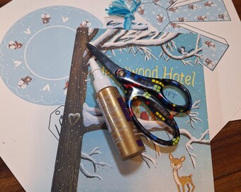 May include: A craft scene featuring a children's book titled "Hollywood Hotel" with a winter illustration, scissors, and a bottle of glue. The scissors have a colorful pattern. There are also cut-out paper shapes with a blue and white pattern.