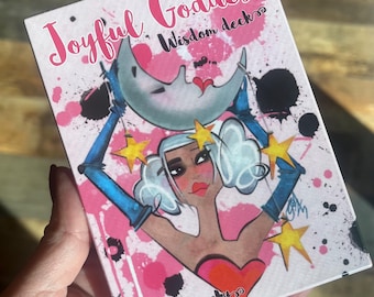 May include: A deck of cards titled "Joyful Goddess Wisdom deck" with a colorful illustration. The artwork features a woman with blue gloves, holding a crescent moon, surrounded by stars and pink paint splatters. The artist's signature is visible.