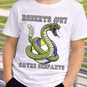 以下が含まれることがあります： 白いTシャツ。緑色のヘビがバレーボールを囲むデザイン。Tシャツには「ROBERTS #27」と「SAYRE SERPANTS」の文字がプリントされています。背景にはフットボールのフィールドが写っています。