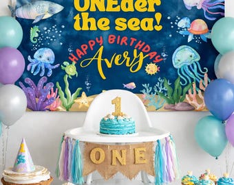 May include: A birthday party scene with an "ONEdeder the sea!" backdrop, balloons, and a high chair decorated with a banner that says "ONE." A blue cake with a gold "1" sits on the high chair. Cupcakes and treats are on the table.