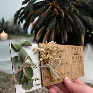 May include: Two wooden save the date cards with a rustic design. The card on the left is white with green eucalyptus leaves and the text "And we're getting married". The card on the right is brown with the text "Save the Date" and "James & Katherine are getting married at Palm Beach".