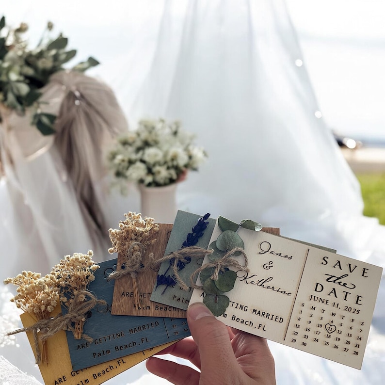 May include: Save the date cards with a rustic wooden design. The cards feature a calendar with the date June 11, 2025, highlighted. The cards are decorated with dried flowers and greenery.