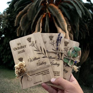 Op de afbeelding: Houten huwelijksuitnodigingen met bloemen- en botanische ontwerpen. De uitnodigingen zijn gegraveerd met de datum "10 oktober 2026" en de namen "William en Audrey". Elke uitnodiging is versierd met gedroogde bloemen en vastgebonden met touw.