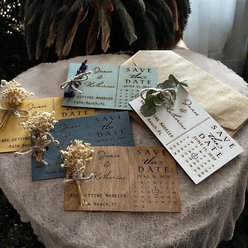 May include: Set of six wooden save the date cards with a calendar design. The cards are decorated with dried flowers and greenery. The text on the cards reads "Save the Date" and includes the names "James & Katherine" and the date "June 11, 2025".