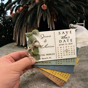 May include: A wooden save the date card with a calendar design. The card is decorated with a eucalyptus sprig and a heart. The text on the card reads "Save the Date" and "June 11, 2025".
