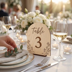 May include: An arched light wood table number sign featuring the text Table 3 in a dark script font, accented with delicate black line art of botanical sprigs on both sides, standing upright on a small rectangular wooden base on a table.
