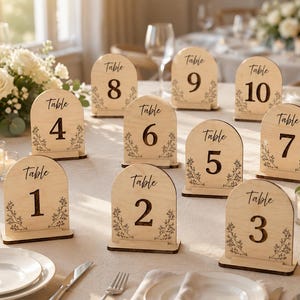 May include: A set of ten arched wooden table number signs, each featuring the word Table followed by a large numeral from 1 to 10. The light-colored wood displays a natural grain and is decorated with delicate black line art of floral sprigs.