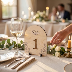 May include: An arched light wood table number sign featuring the word Table and the number 1 in a dark script font, decorated with black line art of botanical sprigs on the sides, standing upright on a small dark base on a table.