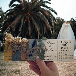 May include: A set of wooden save the date cards with dried flowers and greenery. The cards are tied together with twine and have the text "James & Katherine" and "Save the Date June 11, 2025" on them.